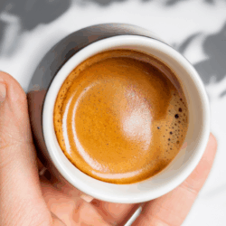An extreme overhead shot looking directly down onto a single, perfectly pulled shot of espresso in a small, matte-black ceramic cup with a clean white rim. The thick, golden-brown crema surface has a defined 'tiger-striping' pattern. A hand holds the cup.