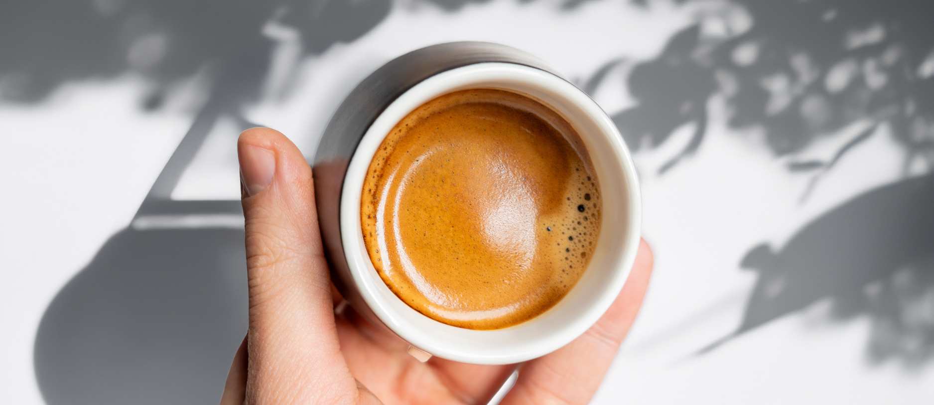 An extreme overhead shot looking directly down onto a single, perfectly pulled shot of espresso in a small, matte-black ceramic cup with a clean white rim. The thick, golden-brown crema surface has a defined 'tiger-striping' pattern. A hand holds the cup.